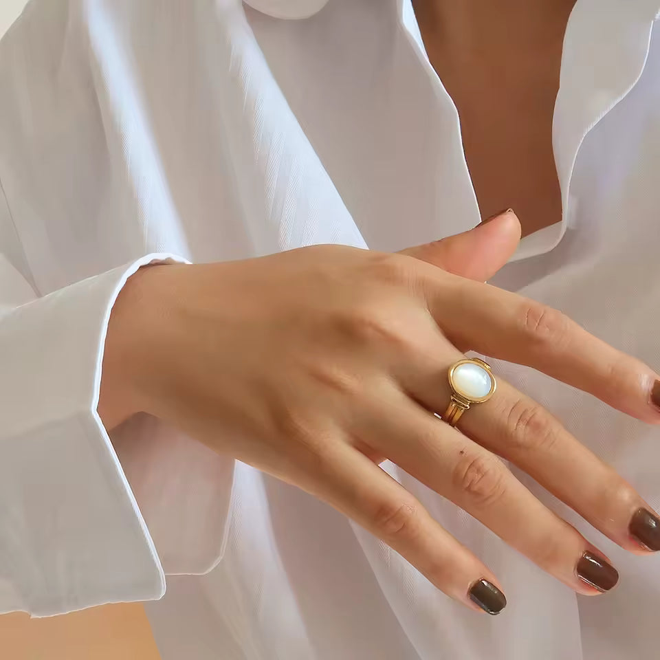 Hand wearing a gold ring with a white stone, against a white shirt background