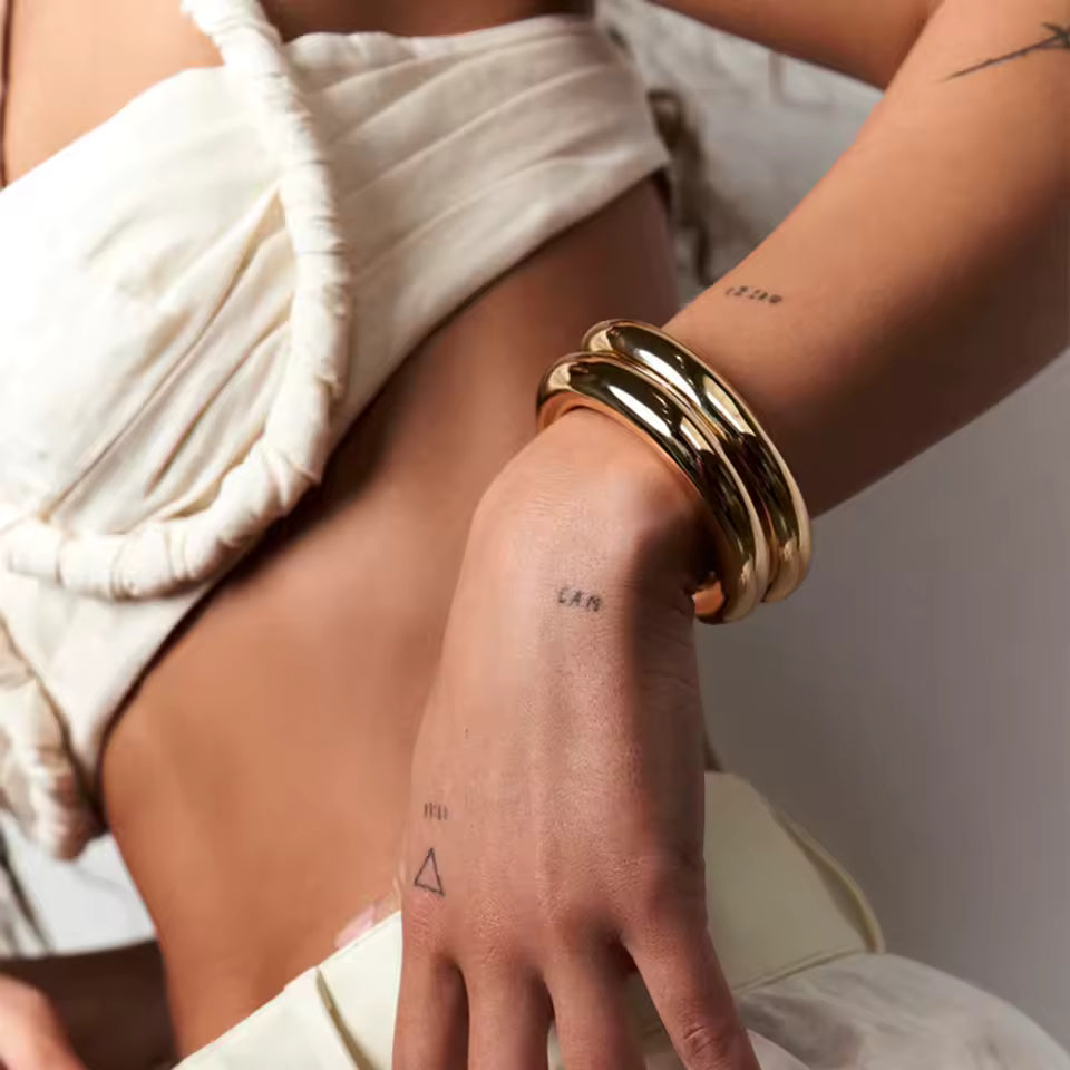 Close-up of a person's arm with gold bracelets and tattoos, wearing a white garment.