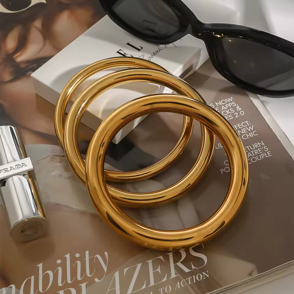 Gold bangles on a magazine with a Prada lip gloss and sunglasses.