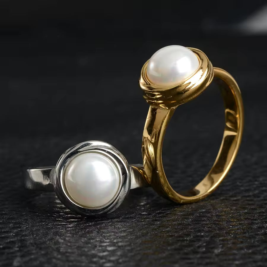 Two pearl rings, one gold and one silver, on a dark textured surface.