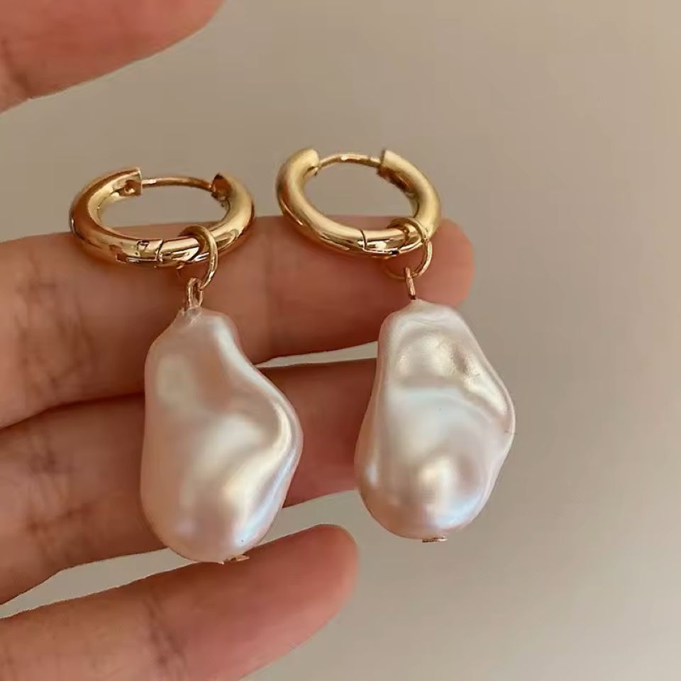 Gold hoop earrings with large pearl drops held in a hand against a neutral background
