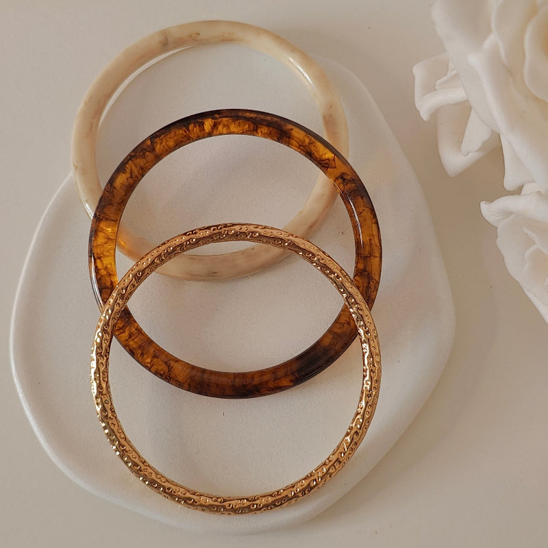 Three bracelets, one off white, one gold and one brown, on a white plate with white background
