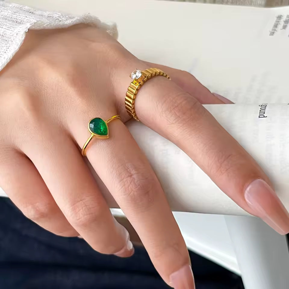 Hand wearing two gold rings with green gemstones on a white surface