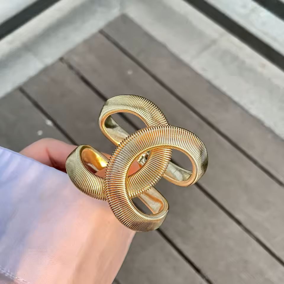 A close-up of a hand wearing a chunky, interlocking gold ring-style cuff bracelet with a ribbed texture, set against a blurred outdoor wooden deck background.