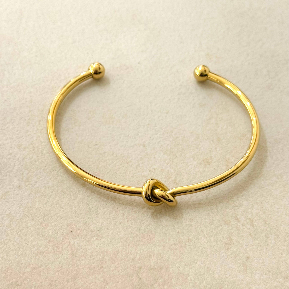 A gold-toned open cuff bracelet with a small knot detail at the center, displayed on a textured beige surface.