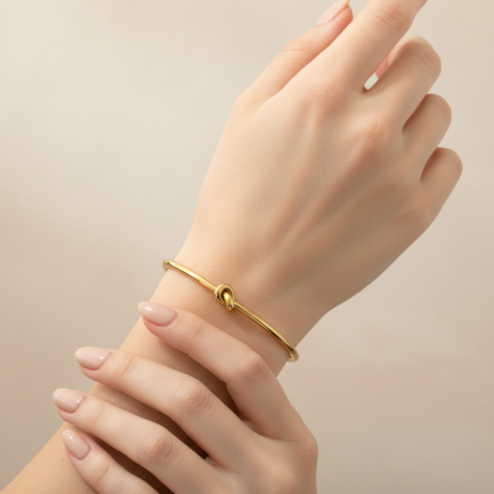 A delicate gold bangle bracelet with a small central knot detail, shown worn on a person's wrist with layered hands.