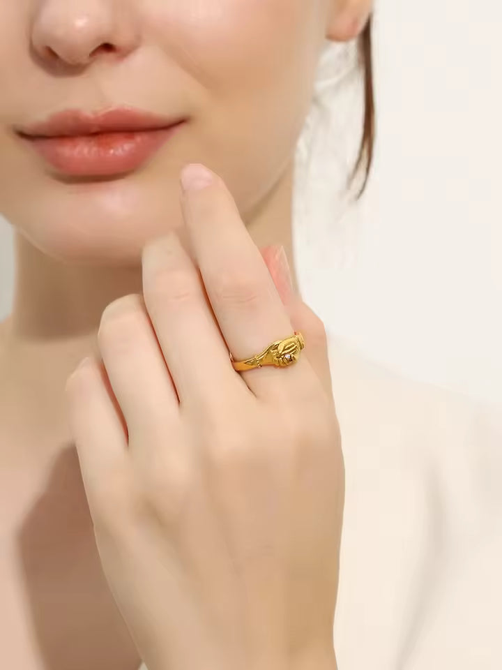 Close-up of a hand wearing a gold ring with hands clasped together design on a neutral background