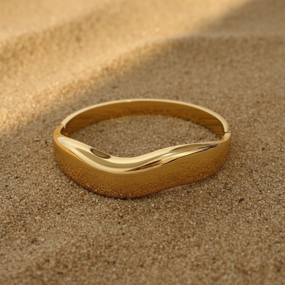 Gold cuff bracelet with wave design laying on the sand