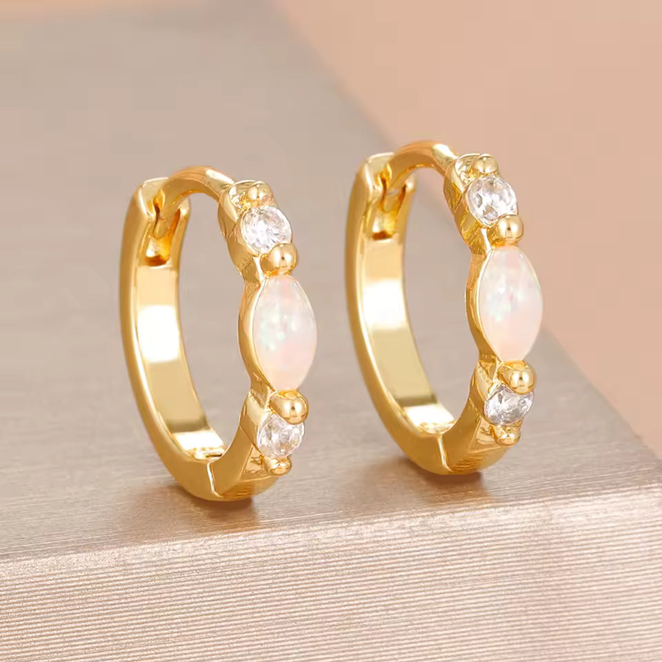 A pair of gold huggie hoop earrings with a central iridescent white opal and small clear cubic zirconia stones, displayed on a textured beige surface.