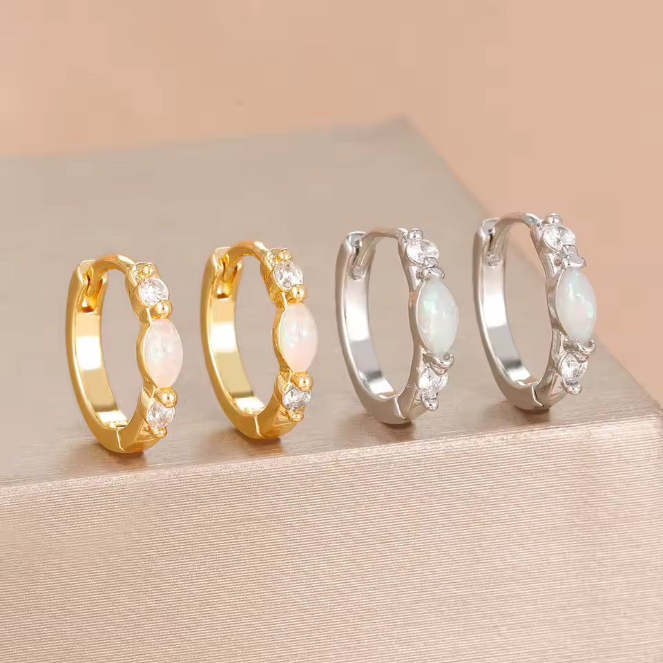 Four small hoop earrings displayed on a beige surface, including two gold-tone and two silver-tone hoops, each with clear rhinestones and an oval milky white center stone.