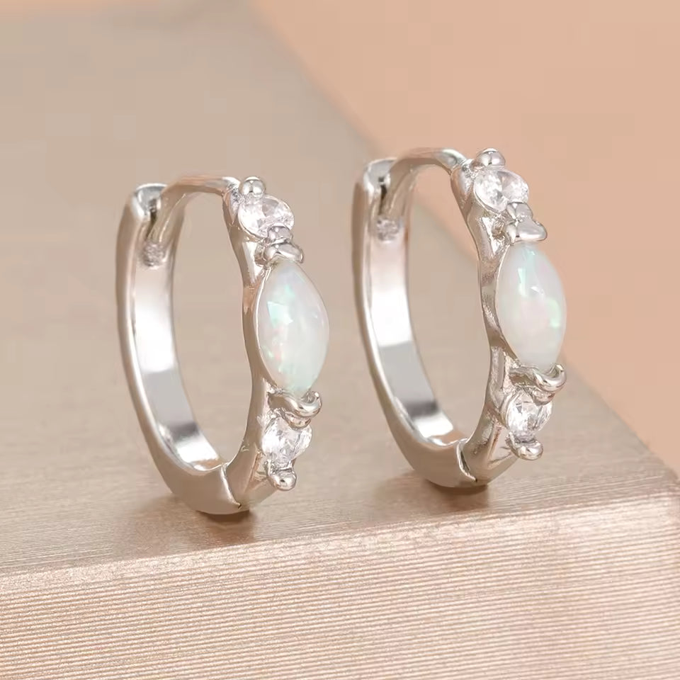 A pair of silver huggie hoop earrings with teardrop-shaped iridescent opal gemstones and small clear crystal accents, displayed on a textured beige surface.