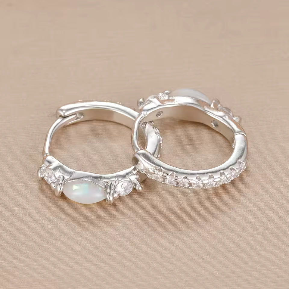 A pair of silver hoop earrings with clear crystal accents and an oval white opal stone, displayed on a textured beige surface.