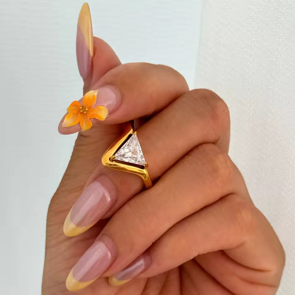 Hand wearing a gold ring with a diamond, small orange flower on nails against a light background