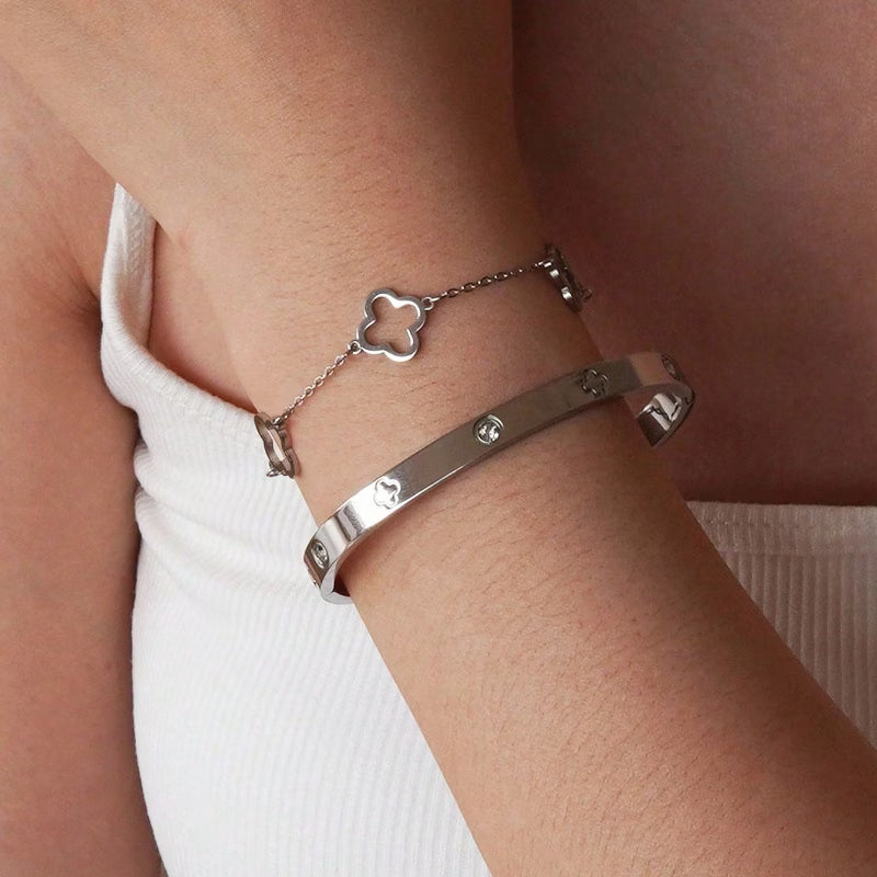 Two silver bracelets worn on a person's wrist, including a bangle with small clear stones and a delicate chain bracelet with a clover motif, shown against skin and a white ribbed top.