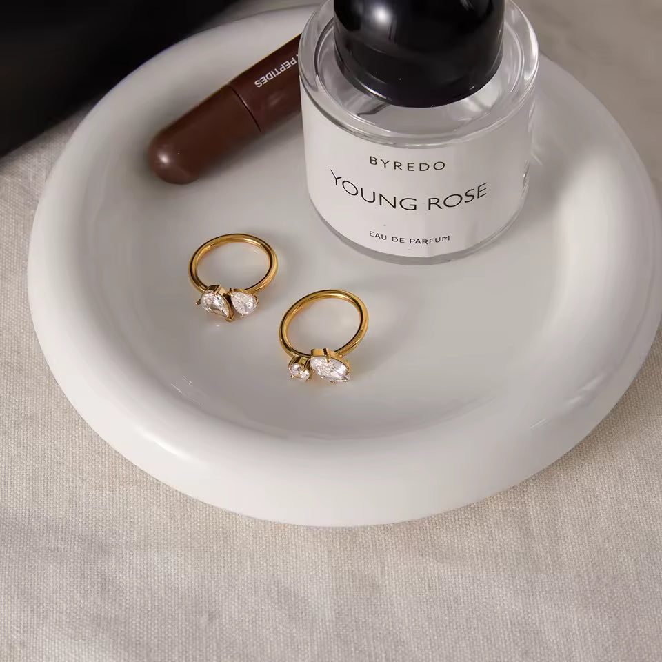 Two gold rings with gemstones on a white dish next to a Byredo Young Rose perfume bottle.
