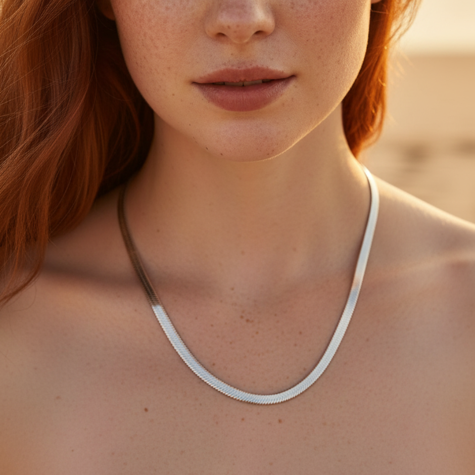 A close-up portrait of a woman with red hair and freckles wearing a thin silver snake chain necklace, set against a warm, softly blurred beach background.