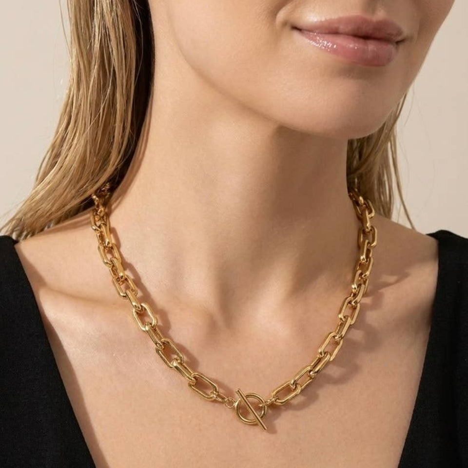A close-up of a model wearing a chunky gold chain necklace with a small interlocking rings clasp, paired with a black V-neck top against a warm beige background.