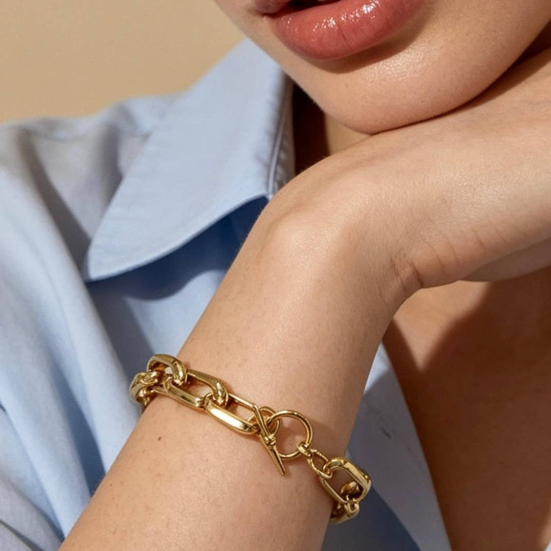 A close-up lifestyle shot of a model wearing a chunky gold chain link bracelet on her wrist, resting her chin on her hand against a light beige background.