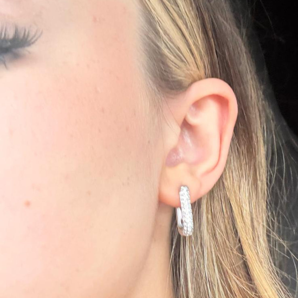 A close-up side profile of a woman's ear wearing a small silver hoop earring with clear stones, against dark hair and a black background.