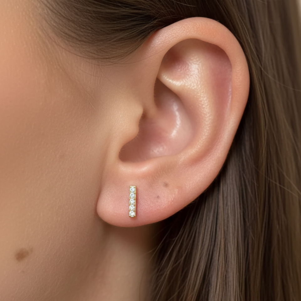 A close-up of a model's ear wearing a small vertical bar stud earring set with tiny clear stones in a yellow metal setting.