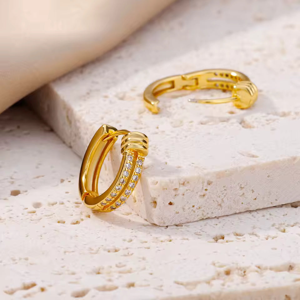 A pair of gold hoop earrings with a row of small clear stones on one earring, displayed on a textured light beige stone surface.