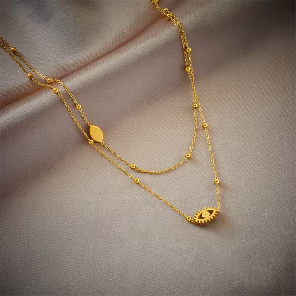 A double-layered gold necklace with a small oval pendant and an evil eye charm, displayed on draped satin fabric.