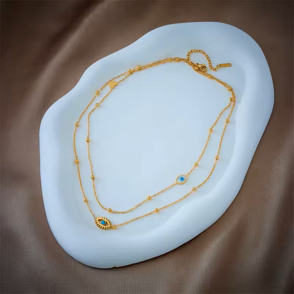 A double-strand gold chain necklace with bead details and an evil eye pendant, displayed on a white organic-shaped tray over a brown satin fabric background.