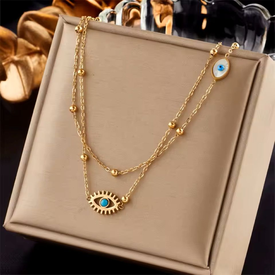 A gold double-layer necklace with evil eye pendants, displayed on a beige jewelry box. The necklace features a textured gold chain with small gold beads and two evil eye charms, one larger on the bottom layer and one smaller oval charm on the upper layer, both detailed with a blue stone in the center.