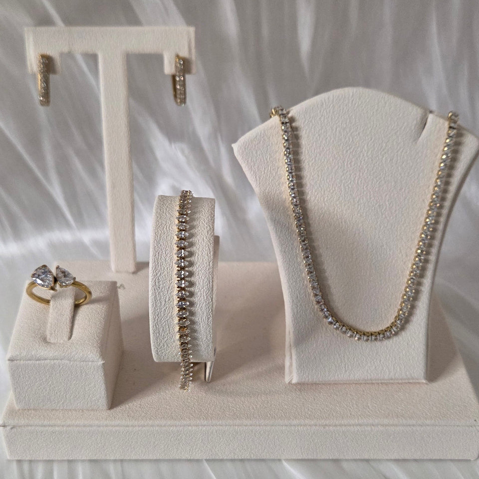 A jewellery set displayed on cream-colored stands, including a gold chain necklace, matching chain bracelet, ring, earrings, and a small charm, set against a draped white satin fabric background.