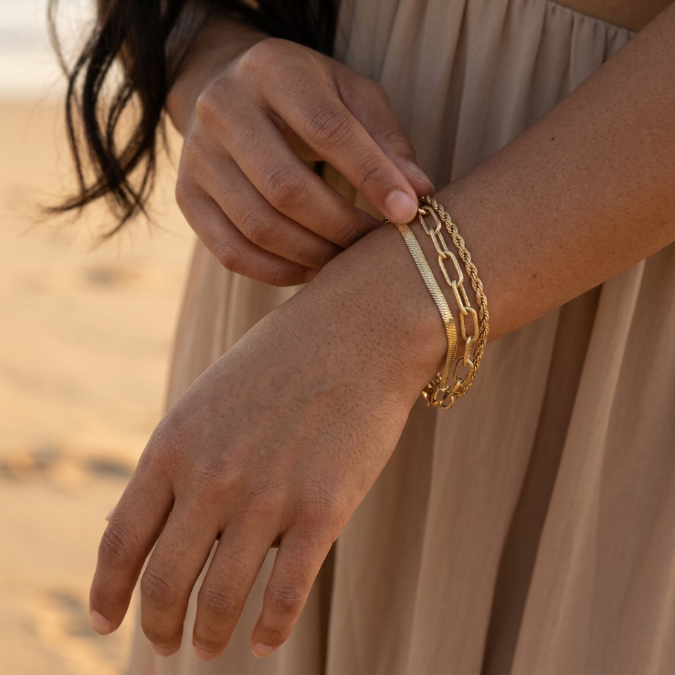 A close-up of a person wearing a stack of three gold chain and bangle bracelets on their wrist, with their other hand adjusting the top bracelet. The background shows a softly blurred sandy beach and beige dress.